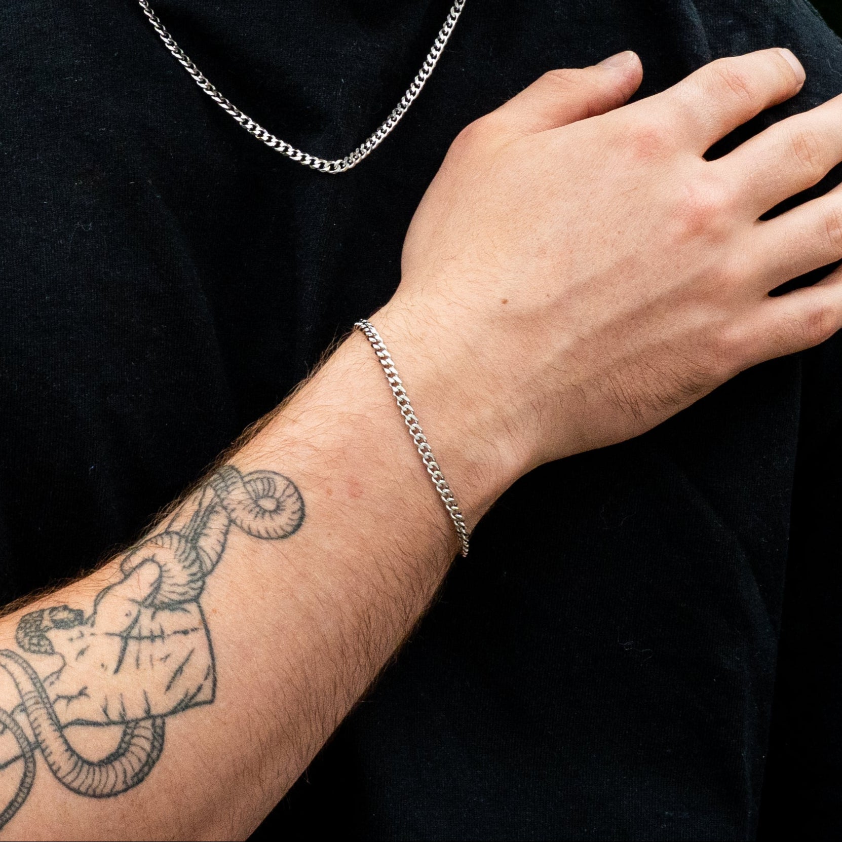 Man wearing a black shirt with a silver chain necklace and bracelet, standing against a green leafy background.